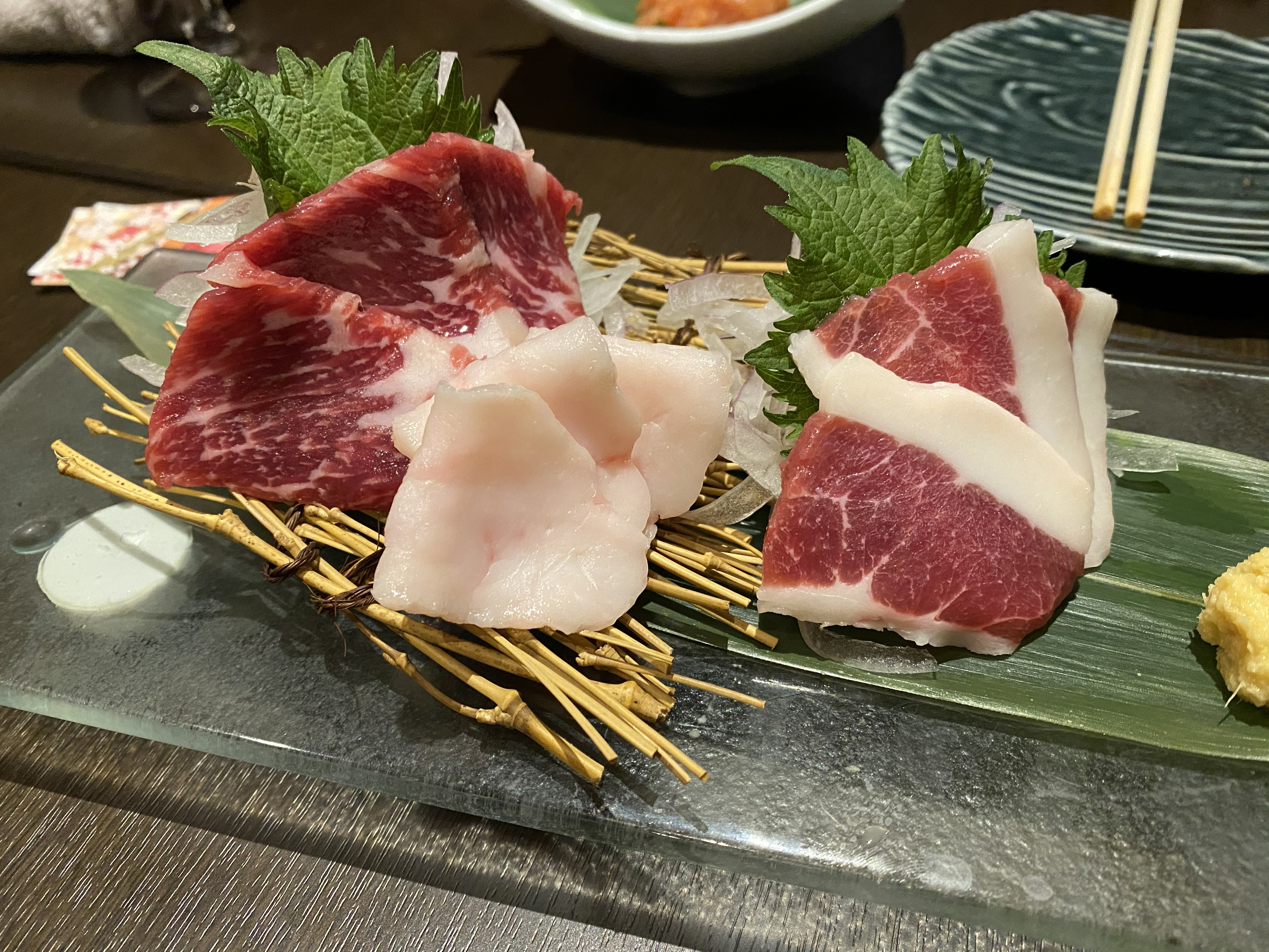 A plate of horse sashimi (basashi) — deep red, well-marbled slices of raw horse meat with shiso leaves and garnish on a glass plate with bamboo matting — a Kyushu specialty served alongside the motsu-nabe