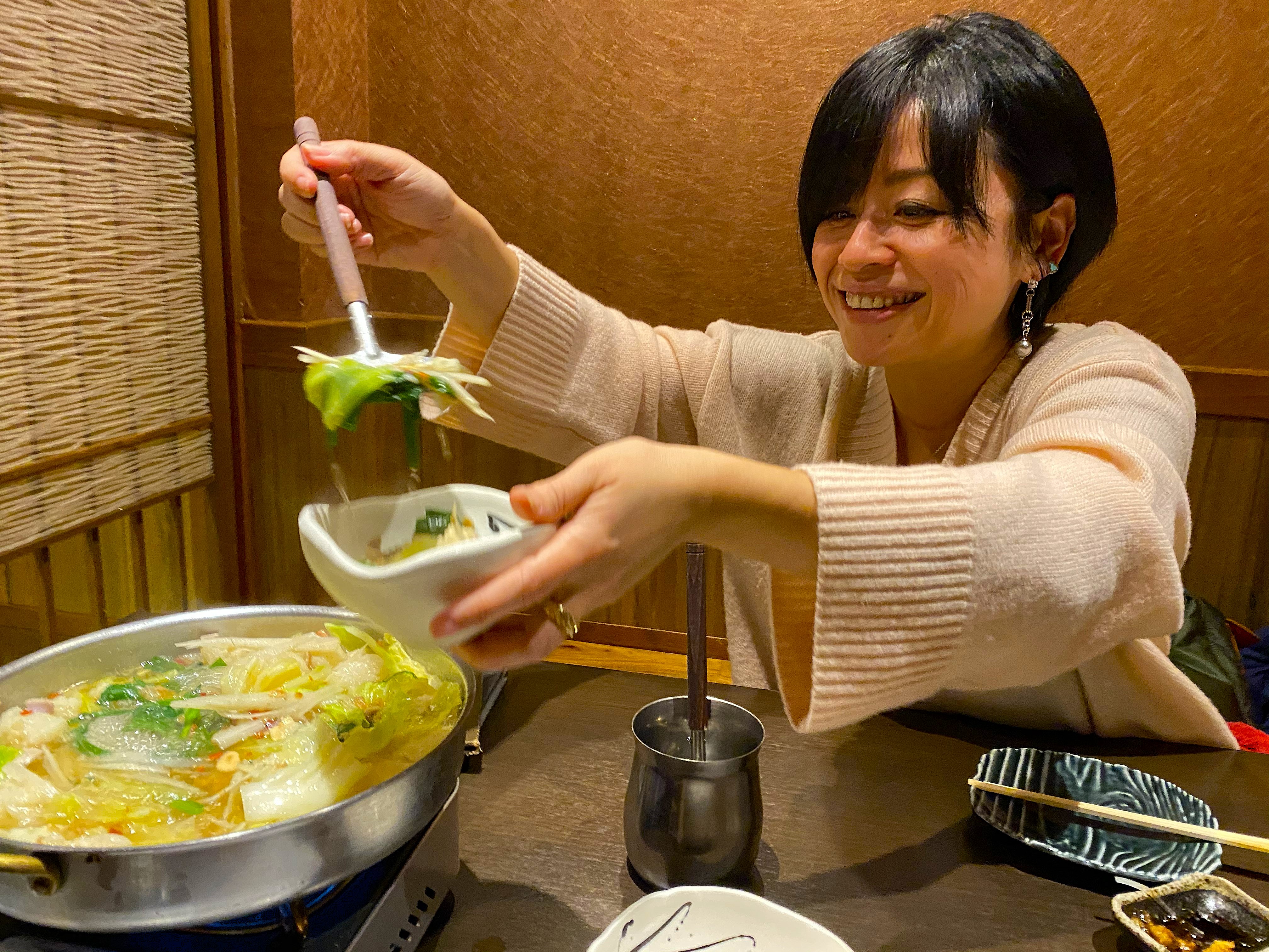 Ayumi serving herself from the motsu-nabe pot at the table — a wide aluminum nabe pan bubbling with cabbage, chives, tofu and offal in a milky broth on the tabletop burner, Ayumi smiling as she ladles into a small bowl