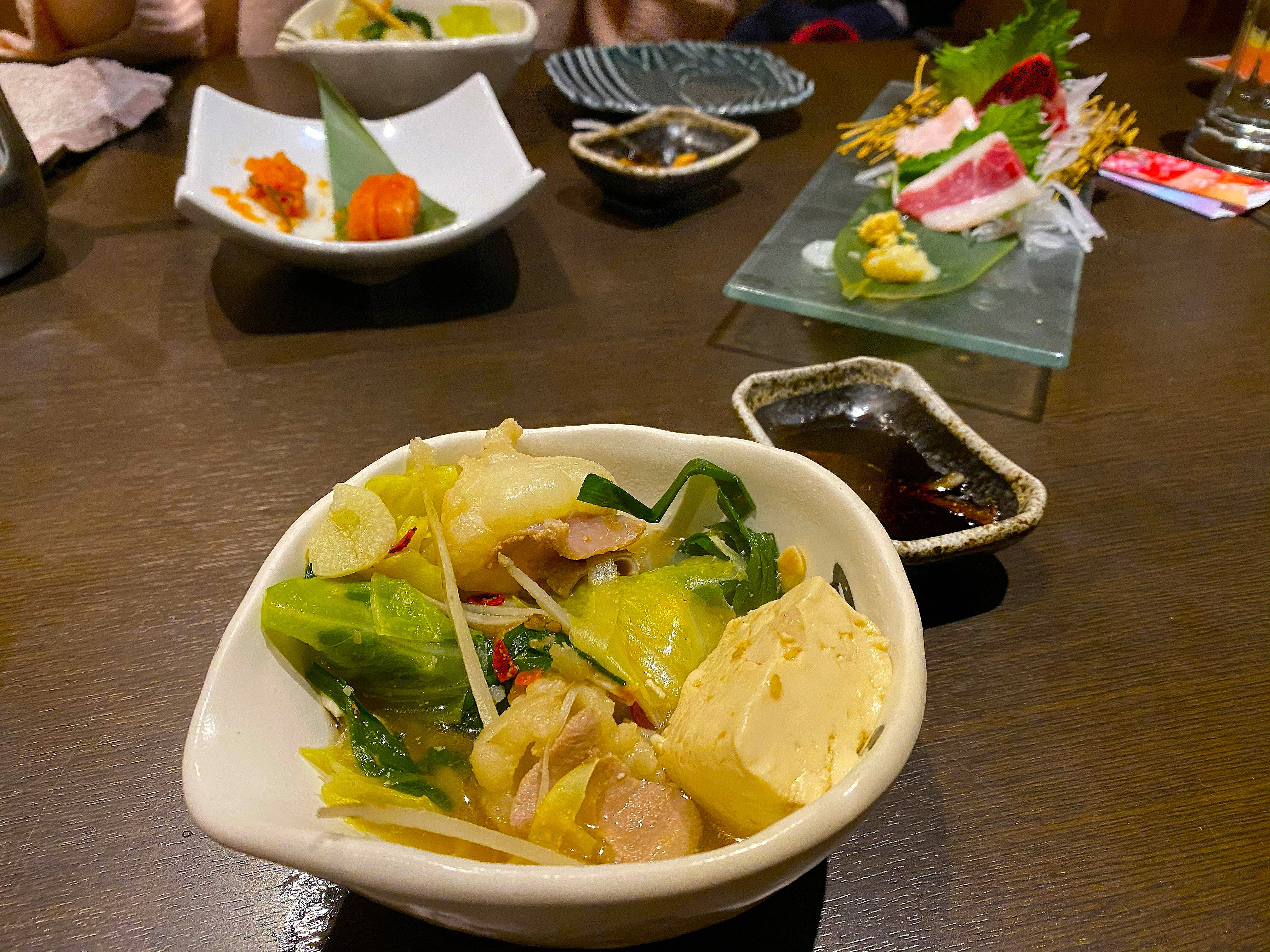 The motsu-nabe table spread — a bowl of nabe contents with cabbage, offal, tofu and vegetables alongside the horse sashimi plate, mentaiko, dipping sauces and side dishes on the restaurant table