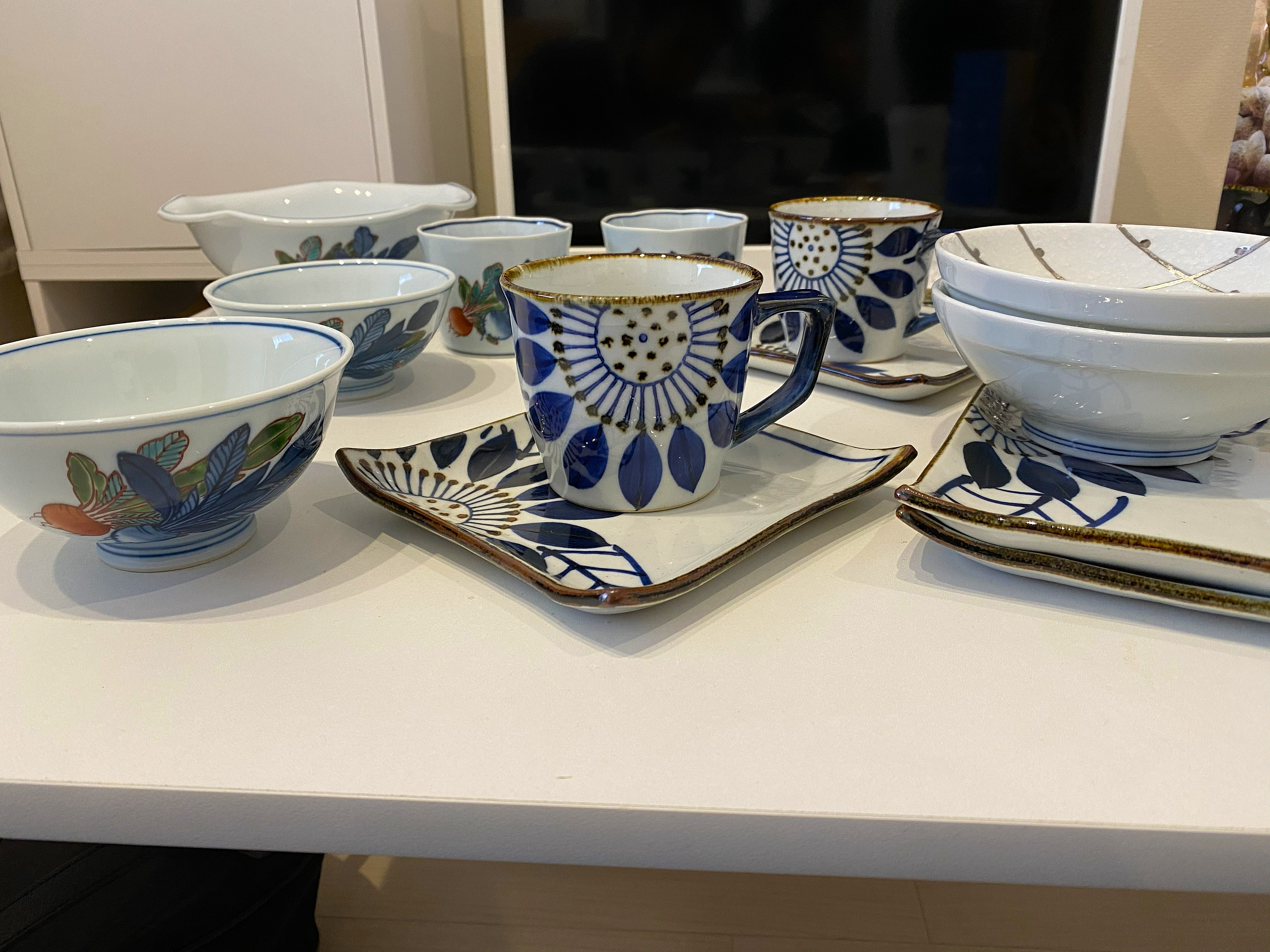 Arita porcelain pieces purchased across multiple shops — bowls with bold leaf and botanical patterns, a coffee mug with a large blue flower motif on a matching square tray, additional plates, all hand-decorated in different kiln styles from Arita Sera and Gen-emon