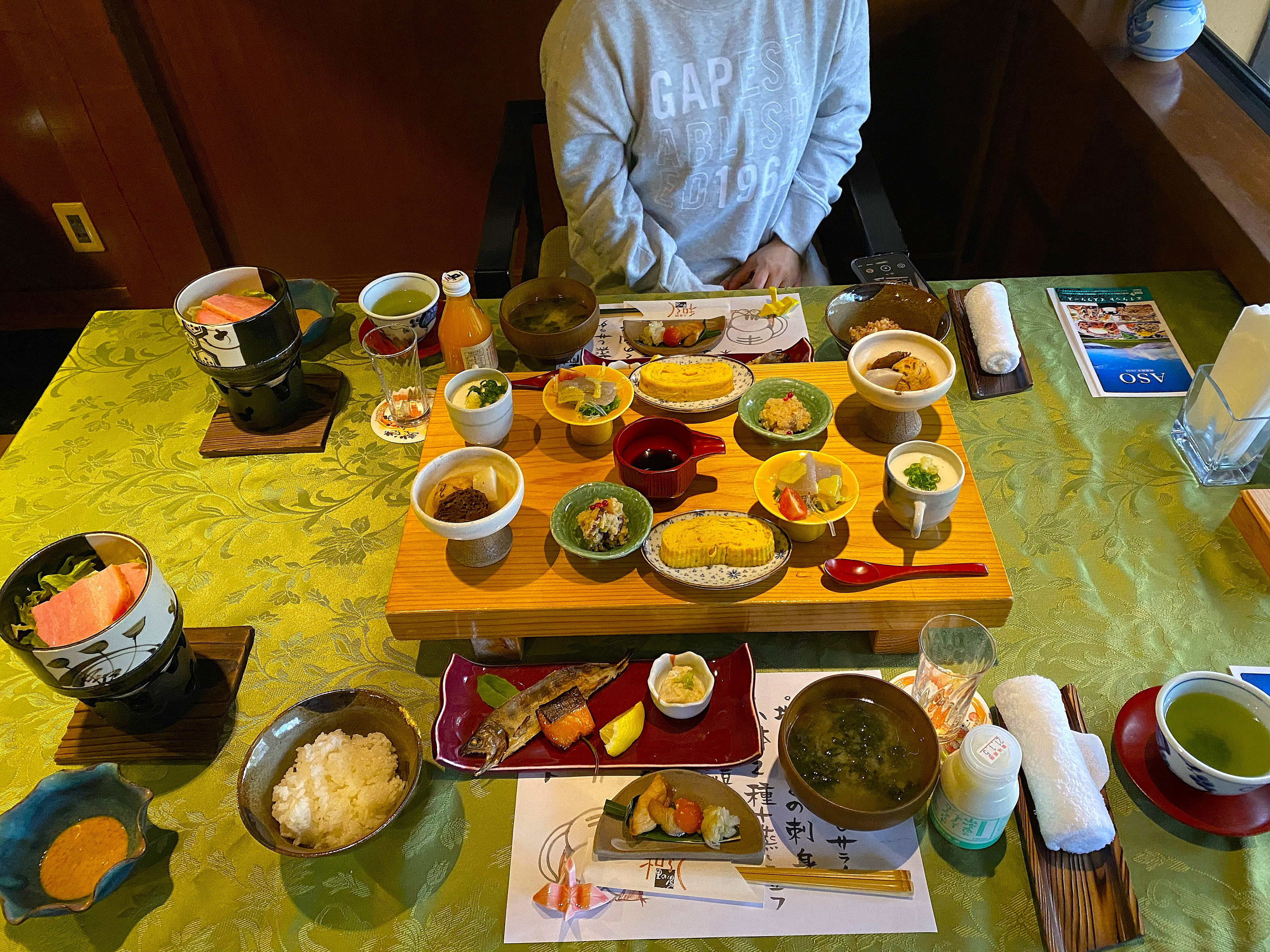 Full Japanese breakfast spread at Satonoyu Waraku ryokan — an elaborate array of small dishes, bowls and plates on a wooden tray, with Ayumi visible behind the table, Kurokawa Onsen guidebook to the side