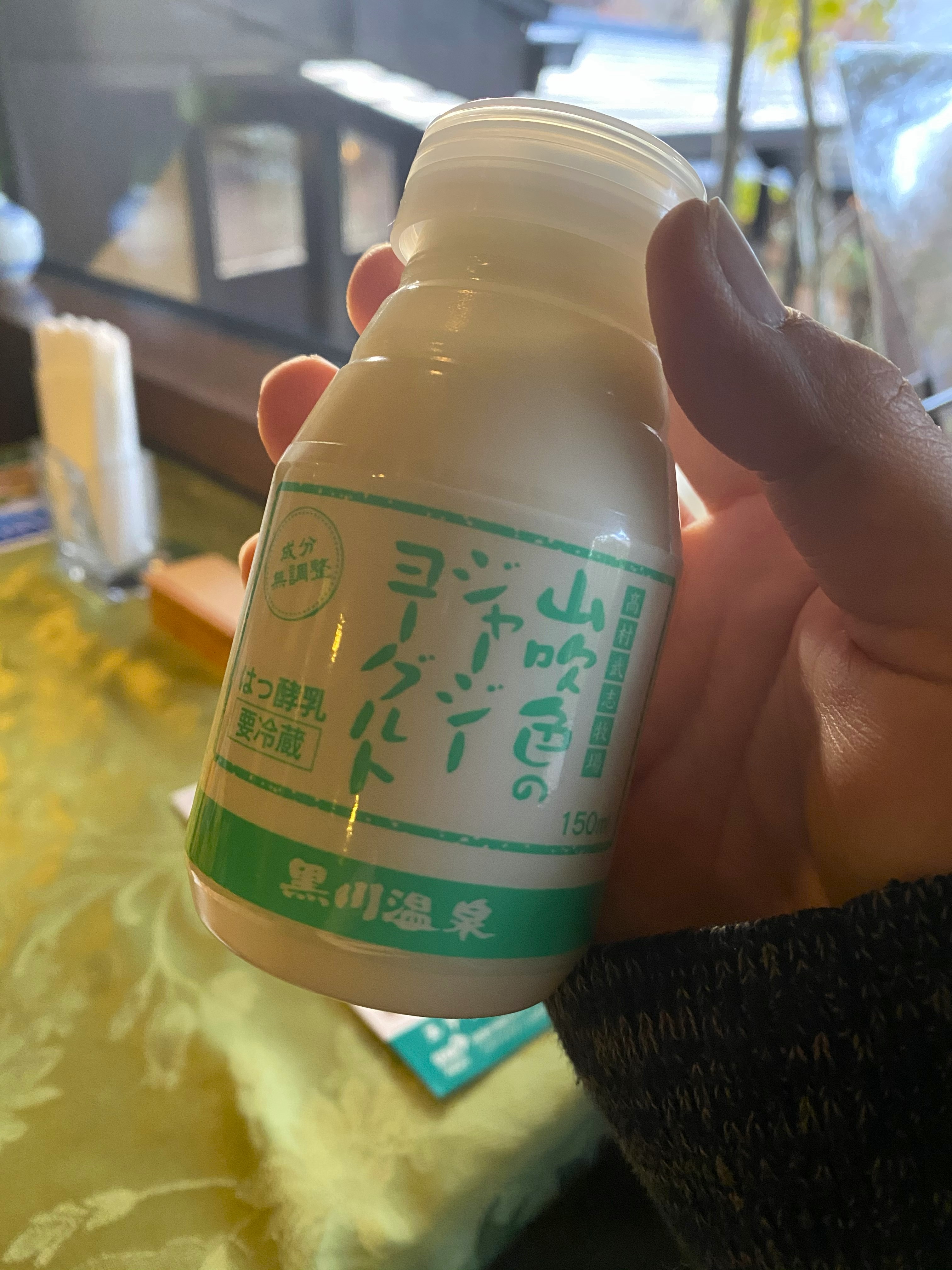 A small bottle of Kurokawa Onsen Jersey yogurt — labeled 山吹色のジャージーヨーグルト, a local specialty made from Jersey cattle milk — held up at the breakfast table