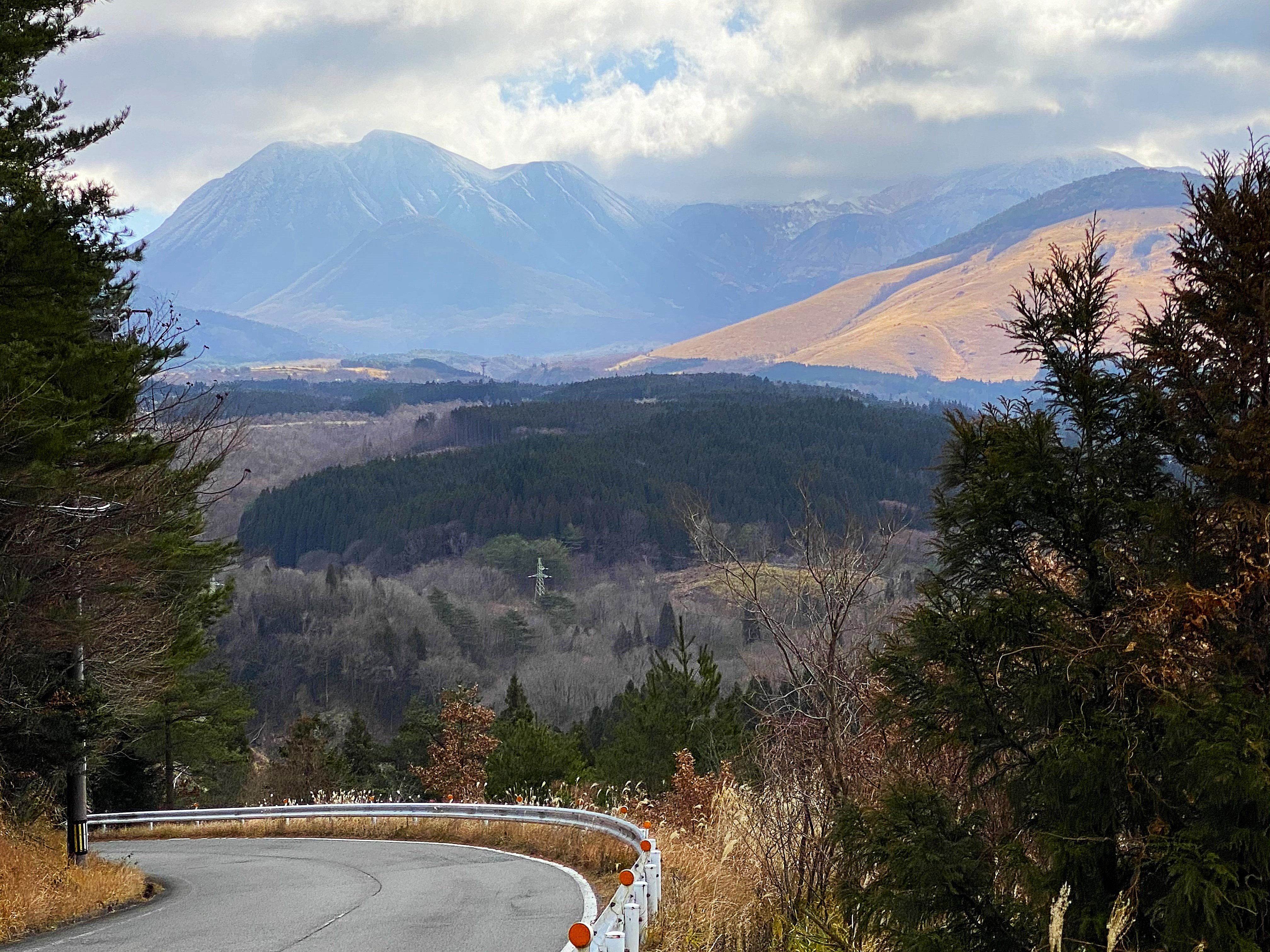 A winding mountain road leaving Kurokawa — snow-dusted peaks rising dramatically above forested ridges in the background, the road curving away below through winter trees, light breaking through overcast cloud