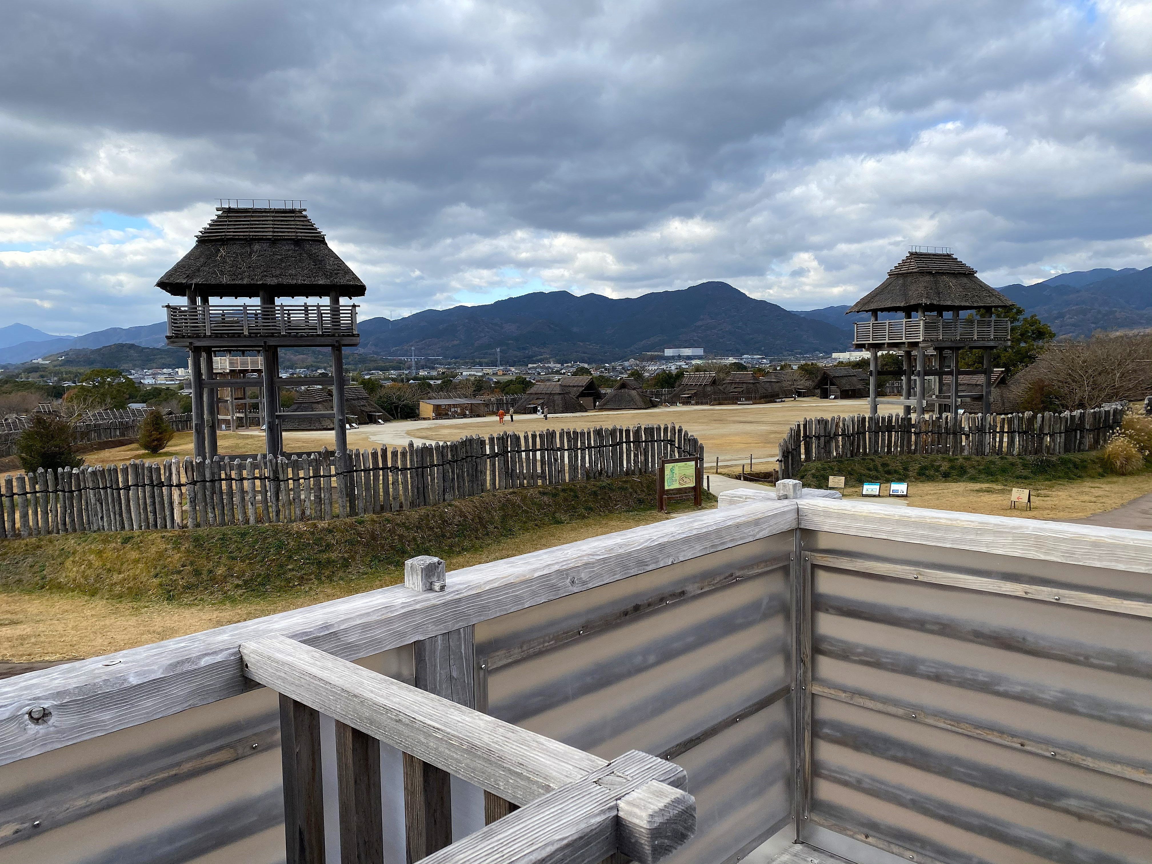 Yoshinogari Historical Park — two reconstructed Yayoi period watchtowers with thatched roofs visible over wooden palisade fencing, the reconstructed settlement spreading behind them, mountains and the modern city visible on the horizon