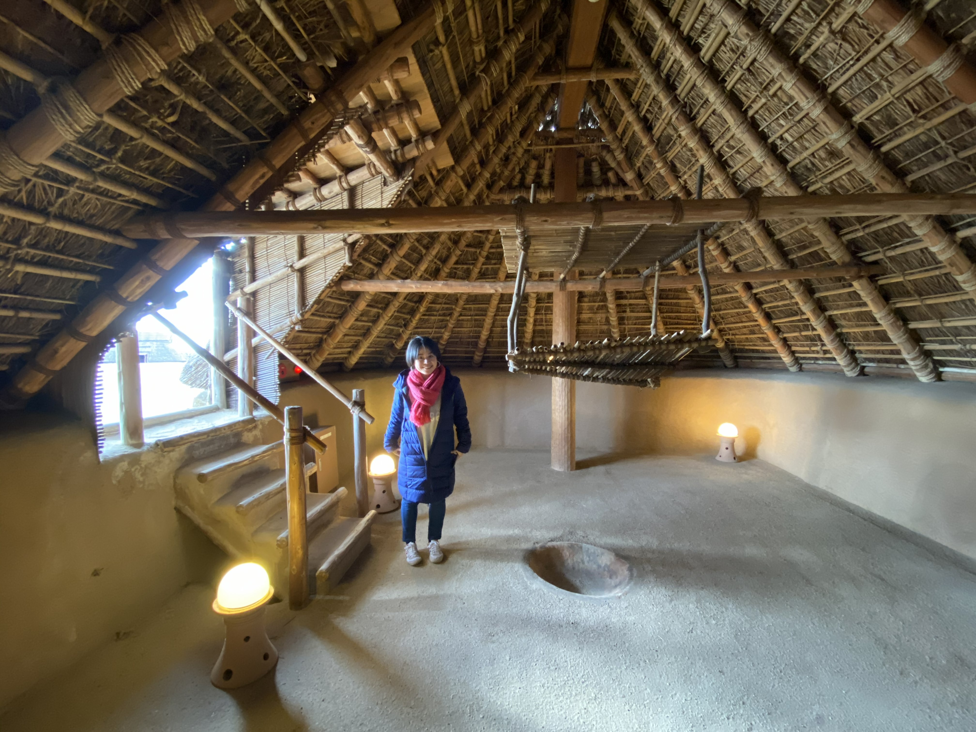 Ayumi standing inside a reconstructed Yayoi period chief's hall at Yoshinogari — the thatched ceiling rising high above, wooden support posts, earthen floor with small floor lamps, a wooden staircase to an upper level