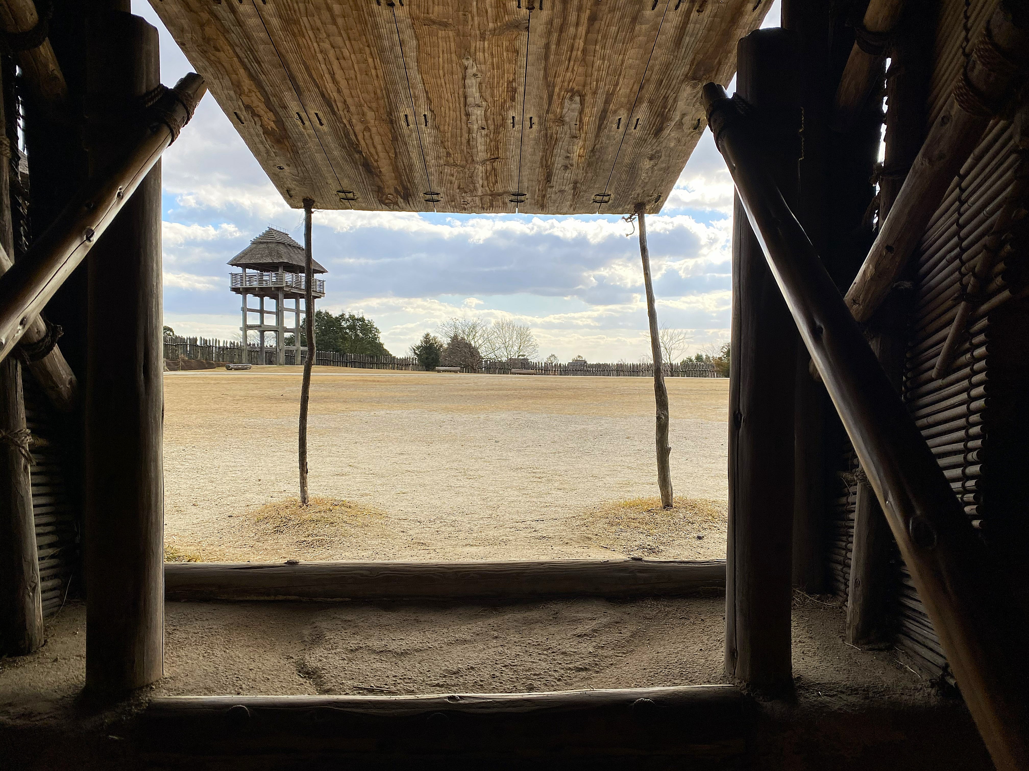 View through the doorway of a reconstructed Yayoi structure at Yoshinogari — wooden frame opening onto the open courtyard, a watchtower visible in the middle distance, sandy grounds stretching to the palisade fence