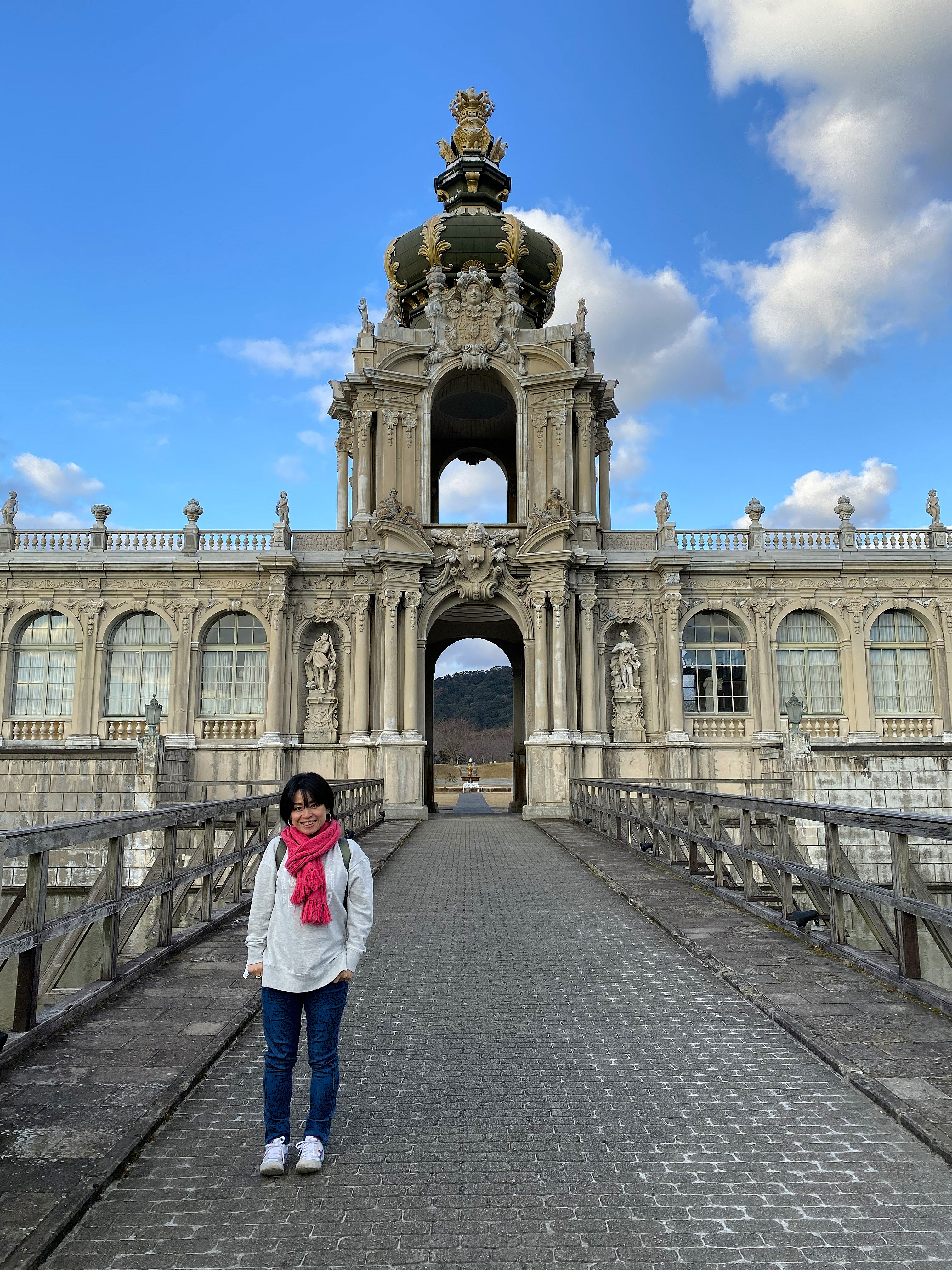 Ayumi standing on the bridge in front of the ornate crown gate tower of the Zwinger replica at Arita Porcelain Park — the baroque tower rises behind her with its gilded crown top, stone arch, and carved figures on either side