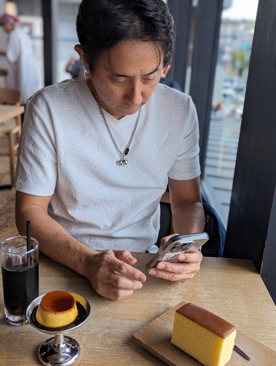 John photographing the castella and custard pudding at Shokado Kashiten — white textured shirt, chain necklace, iced coffee at his side