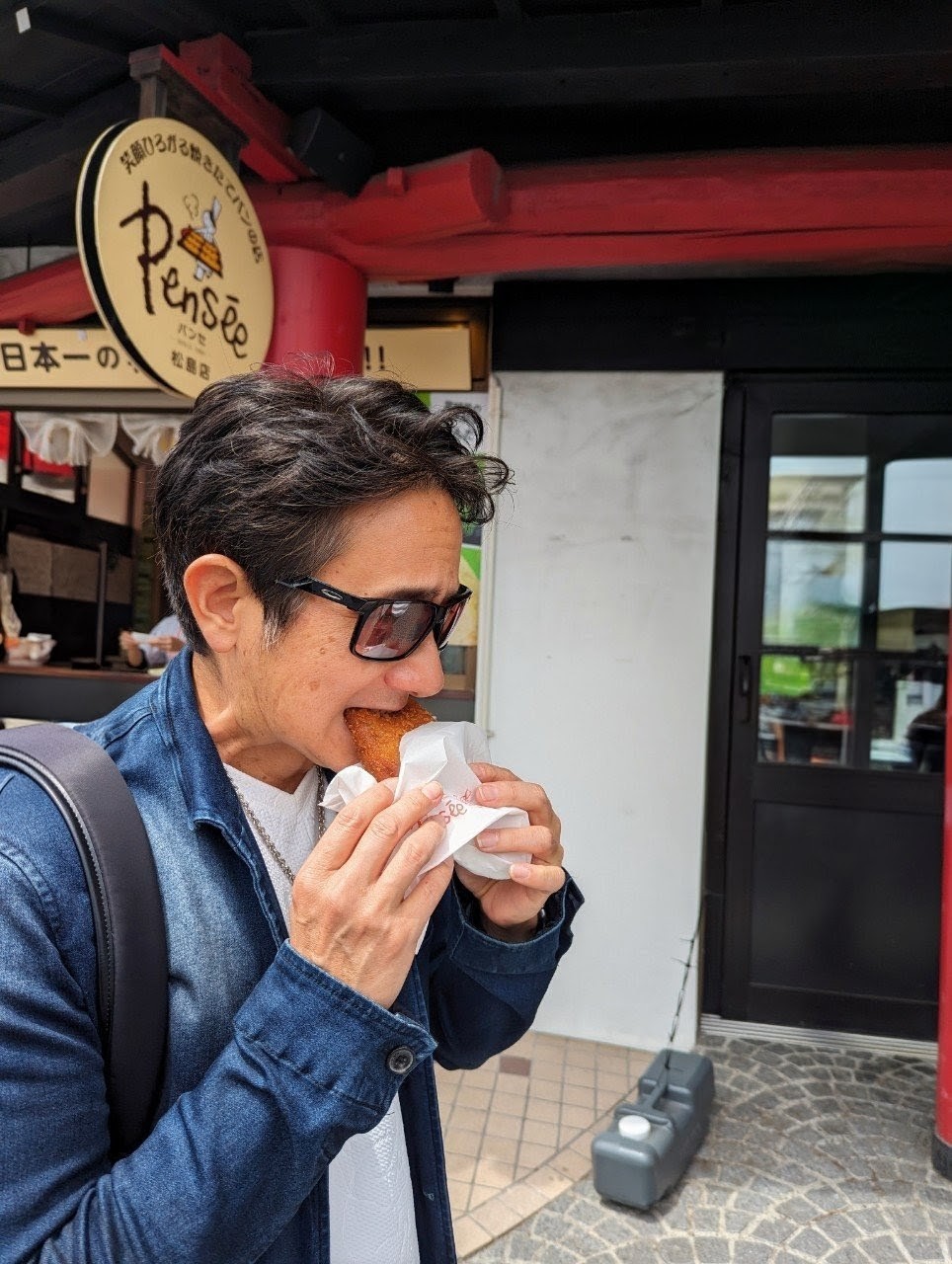 John eating the Pensée kaki curry bread outside the Pensée bakery in Matsushima — denim jacket, sunglasses, the Pensée sign visible behind him