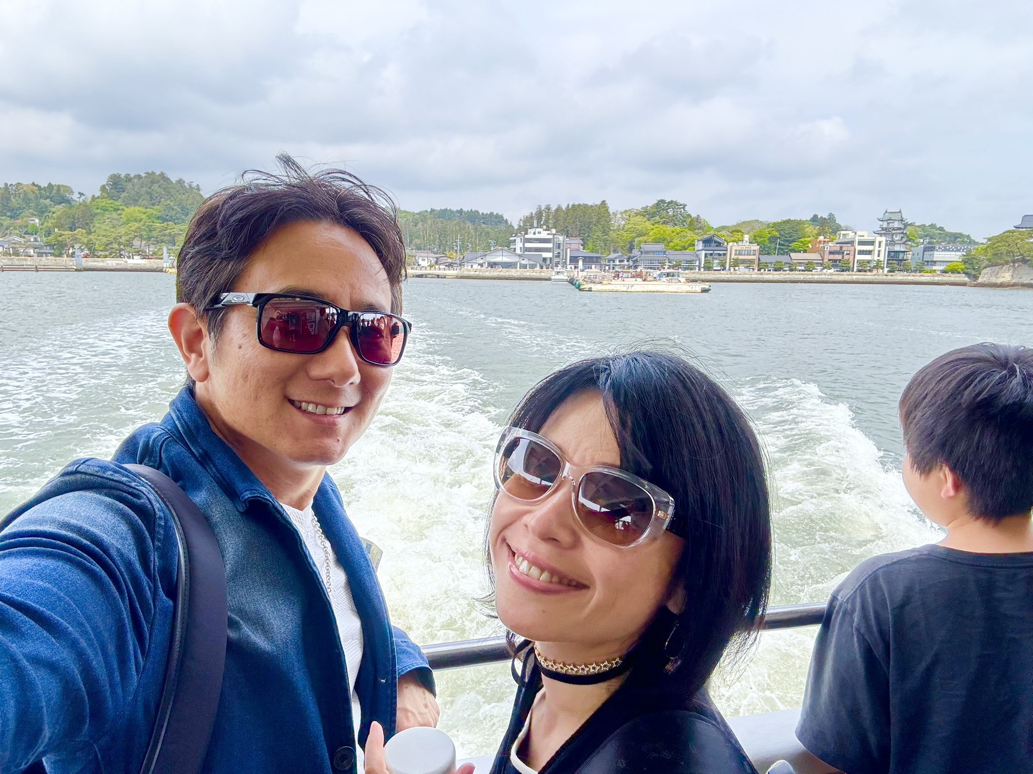 John and Ayumi smiling on the back deck of the Matsushima ferry, the bay and Matsushima waterfront visible behind them