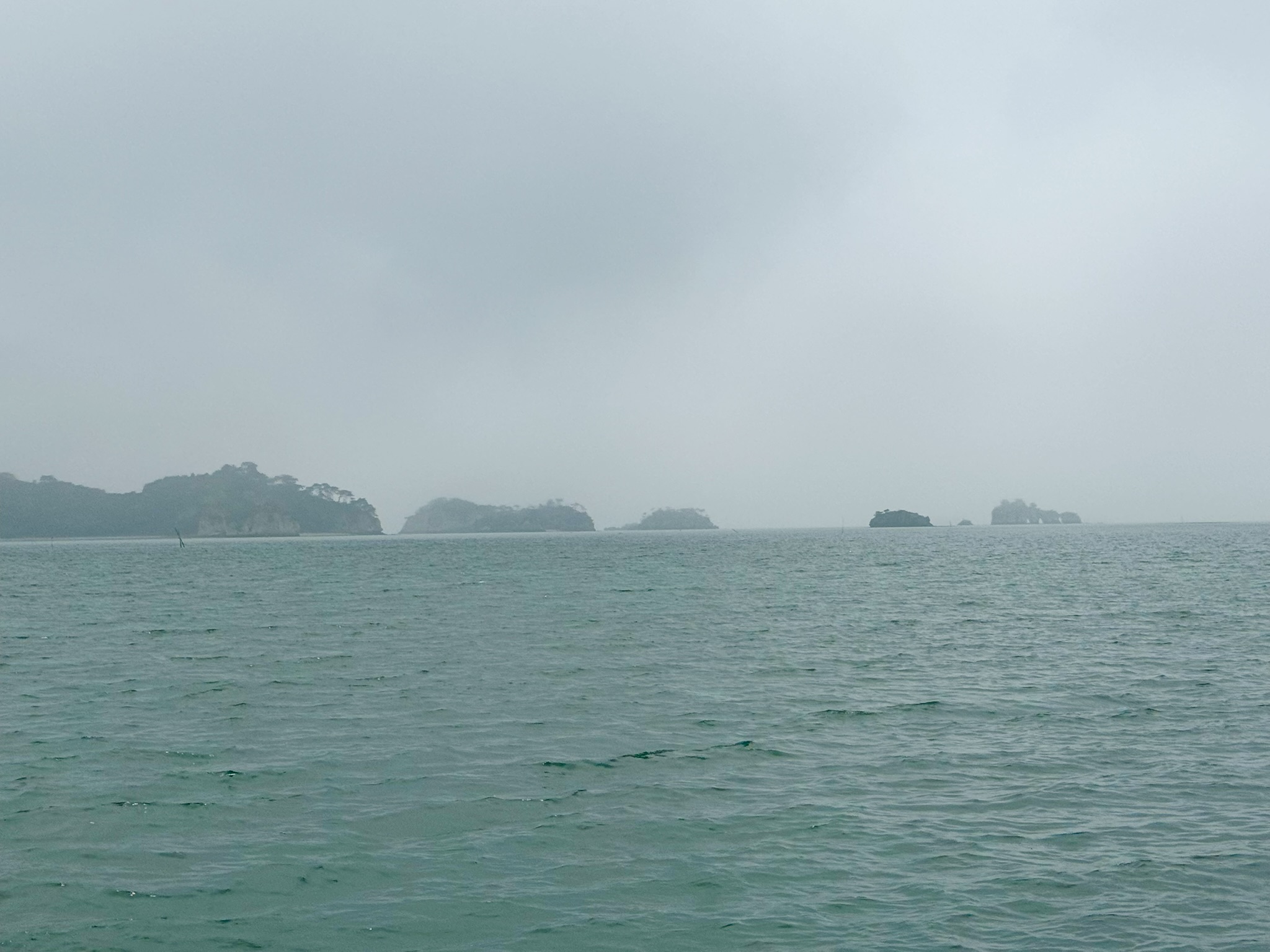 Matsushima Bay from the ferry — pine-covered islands rising from flat grey-green water under a heavy overcast sky, the far islands barely visible through the haze