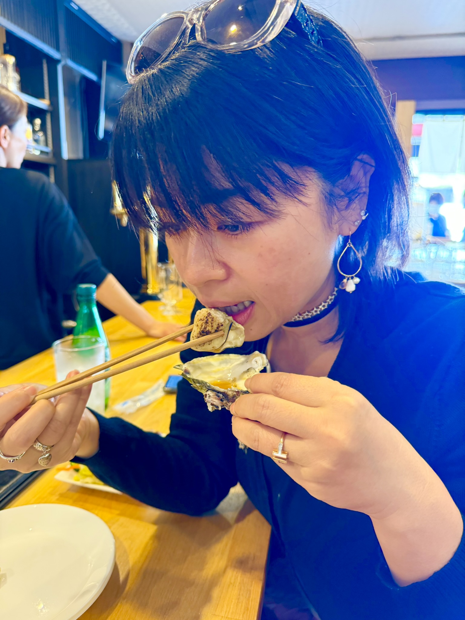 Ayumi eating a grilled Matsushima bay oyster with chopsticks at a restaurant in Matsushima, sunglasses pushed up on her head