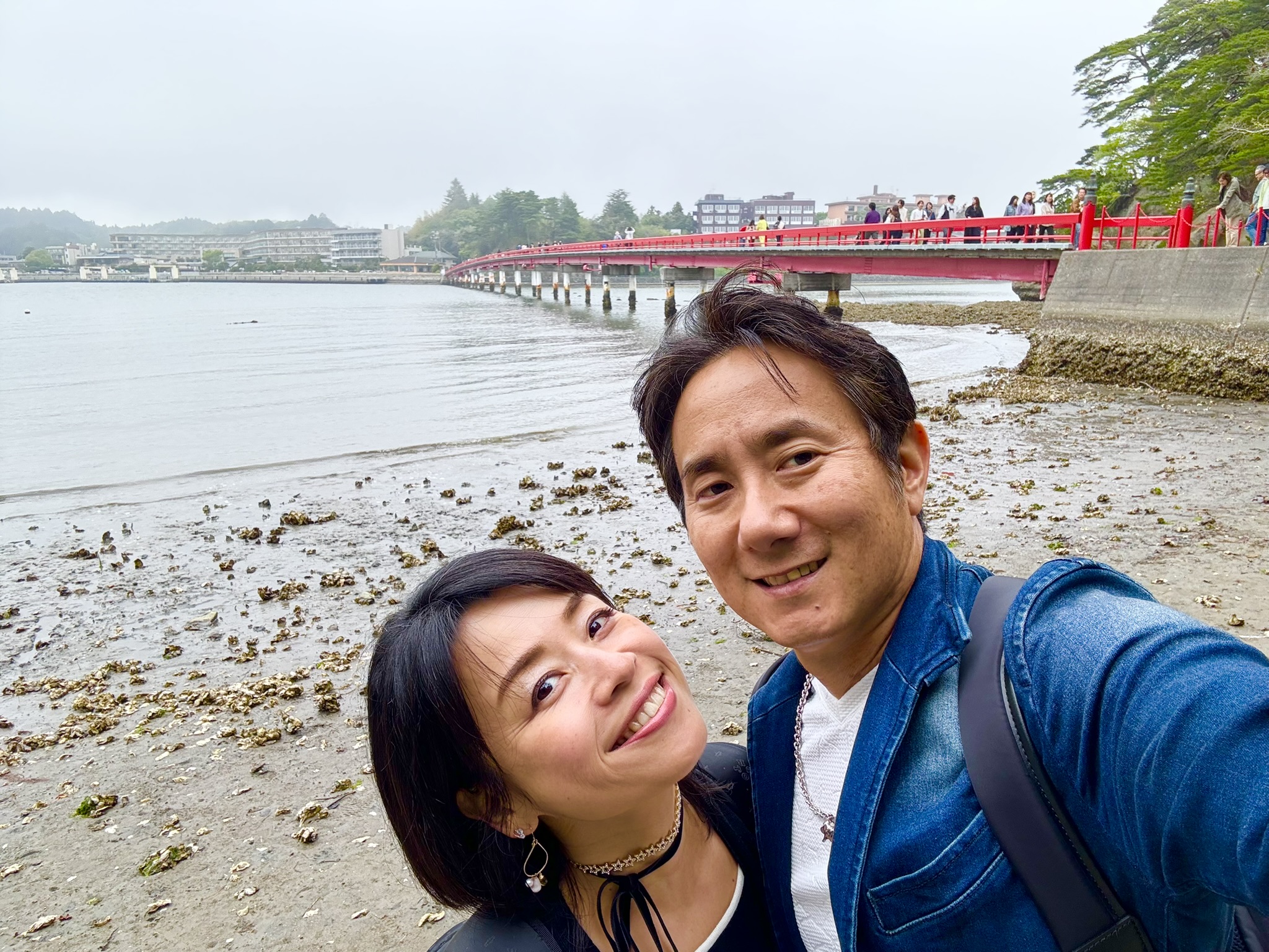 The red Fukuurajima pedestrian bridge at Matsushima viewed from Fukuurajima island — the iron span curving over grey water, backed by pine-covered hills and a hazy sky