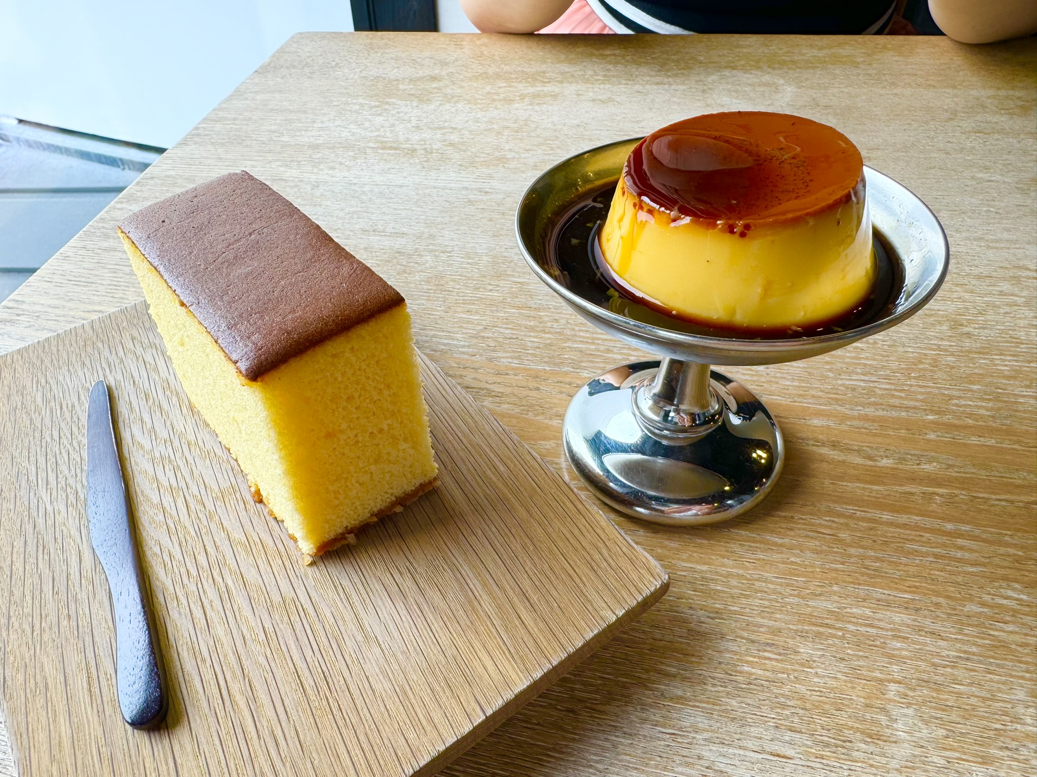 Castella cake on a wooden board and a Japanese custard pudding on a chrome pedestal dish, side by side on a light wood café table at Shokado Kashiten in Matsushima