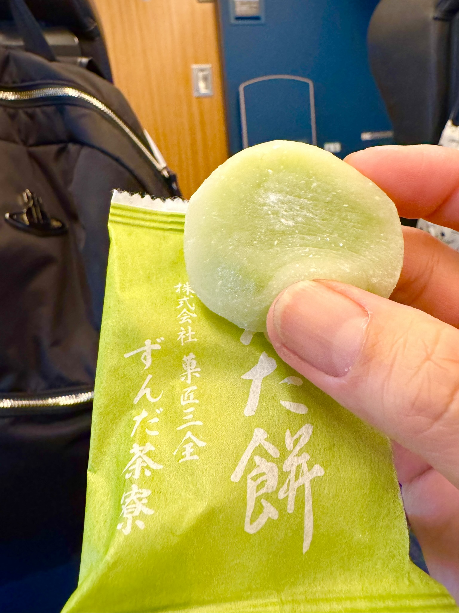 A hand holding a round, pale green zunda mochi in front of a bright green package from Kashosanzen Zunda Chaya — the kanji for zunda mochi visible on the packaging