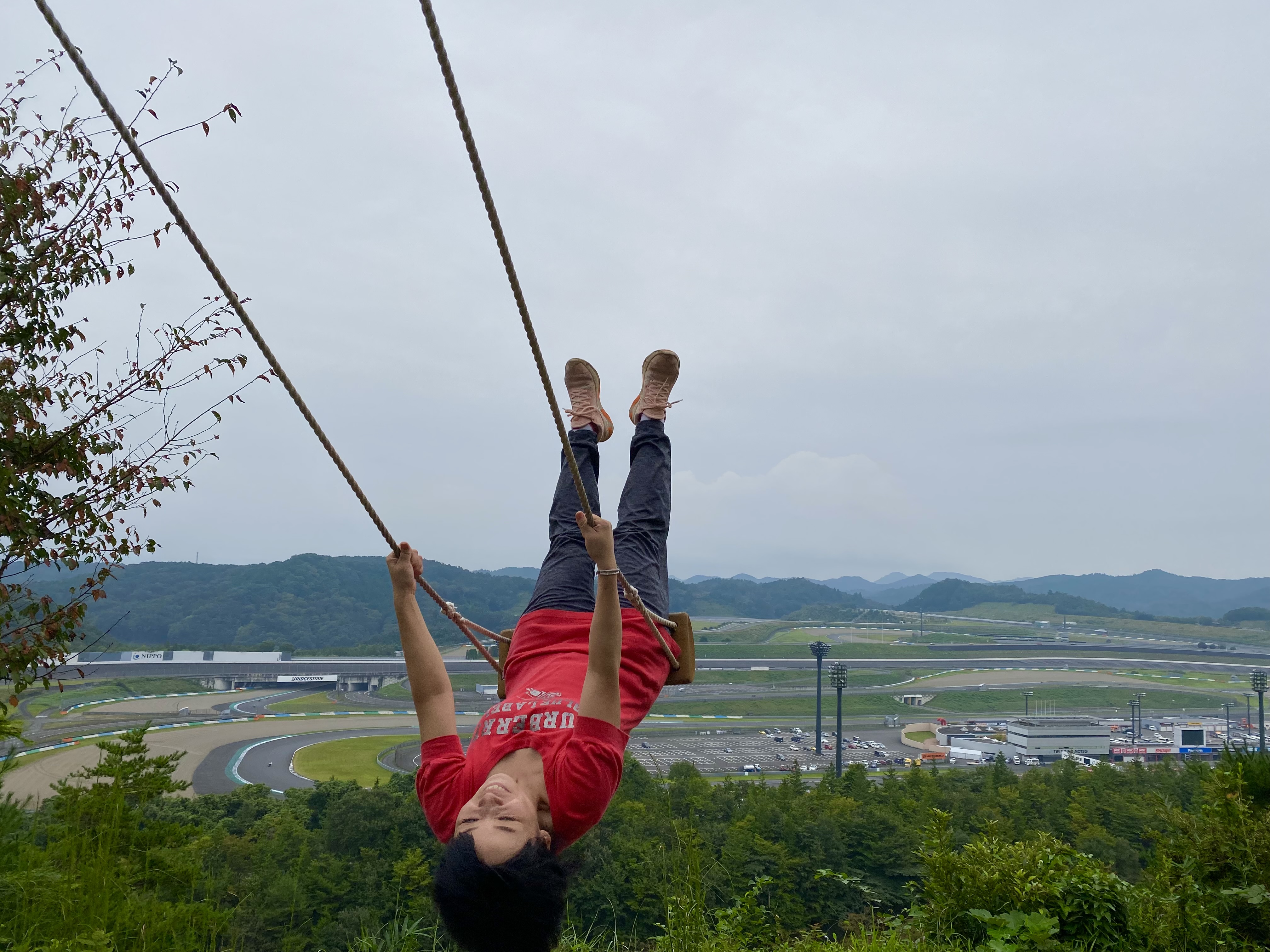 Person on a giant rope swing at the Musasabi course, swinging out over the hillside with the Twin Ring Motegi race circuit visible below