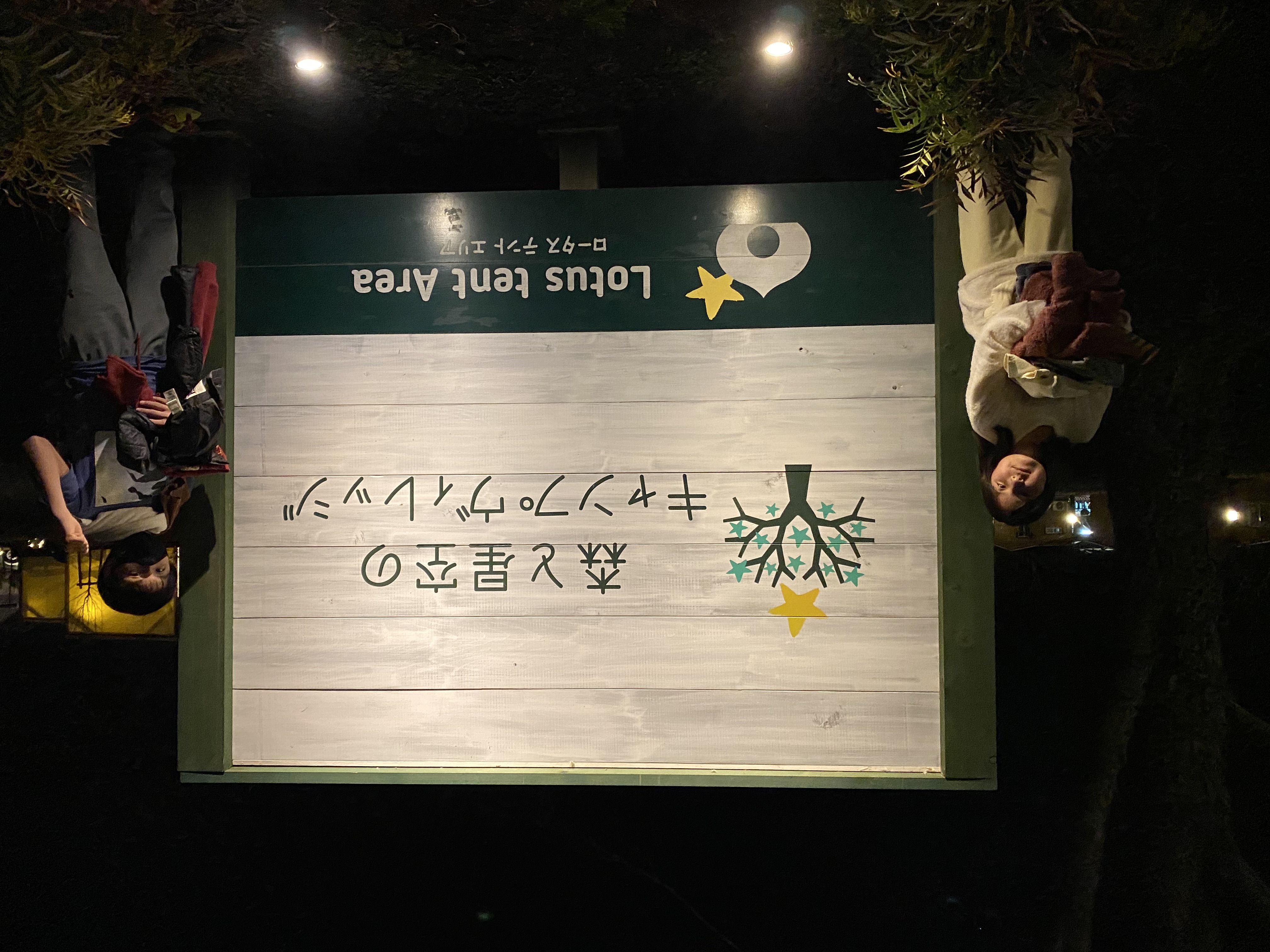 Jace and Maile standing in front of the lit-up Forest and Starry Sky Camp Village sign at night, with the Lotus tent area sign below