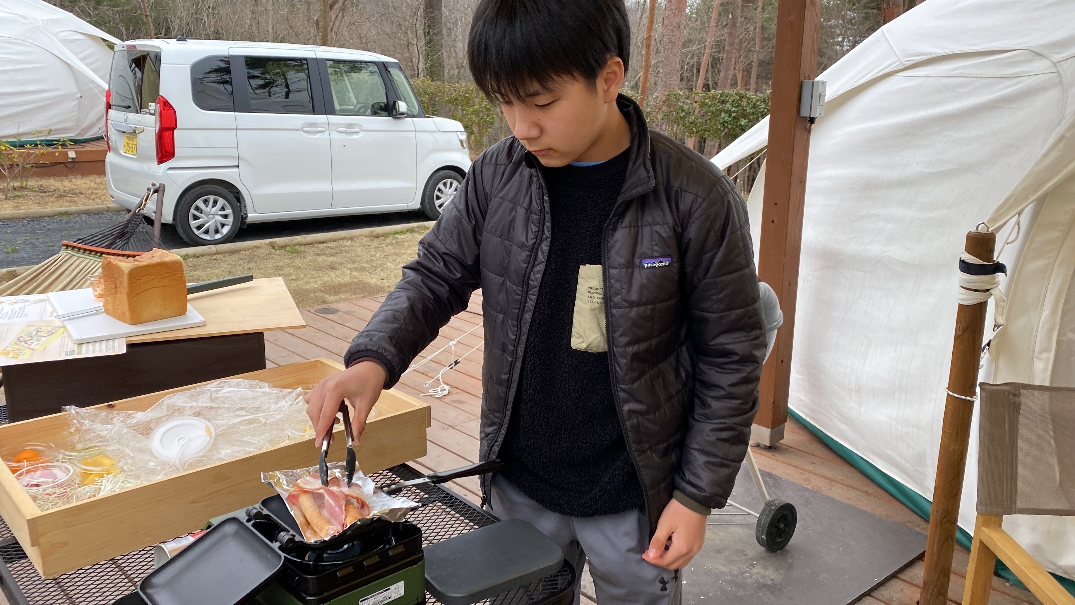 Jace grilling bacon on a portable grill on the deck in the morning with the breakfast spread visible on the table behind him
