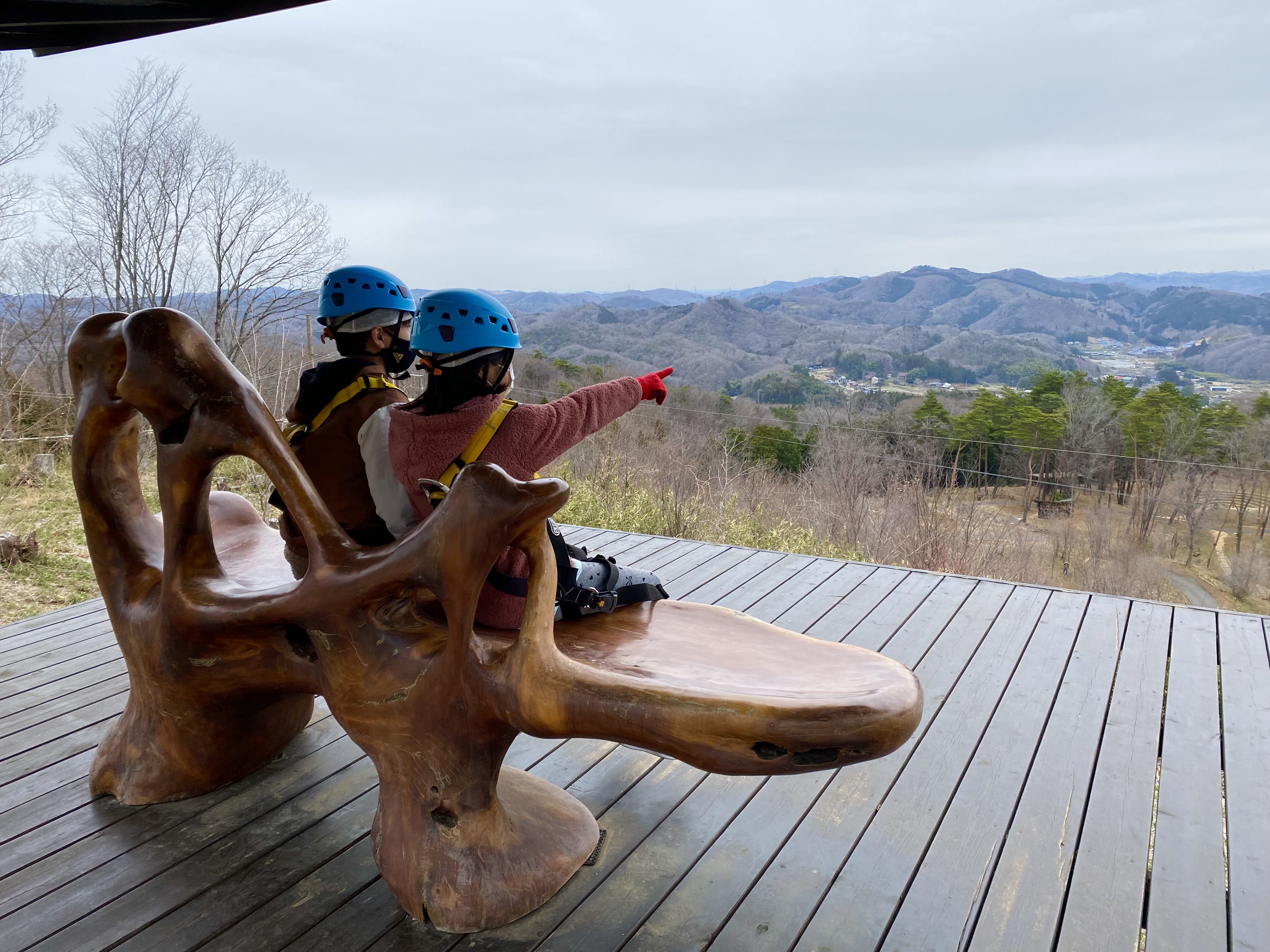 Jace and Maile in blue helmets sitting on a large carved wooden bench at the Musasabi course, pointing at the mountain views beyond