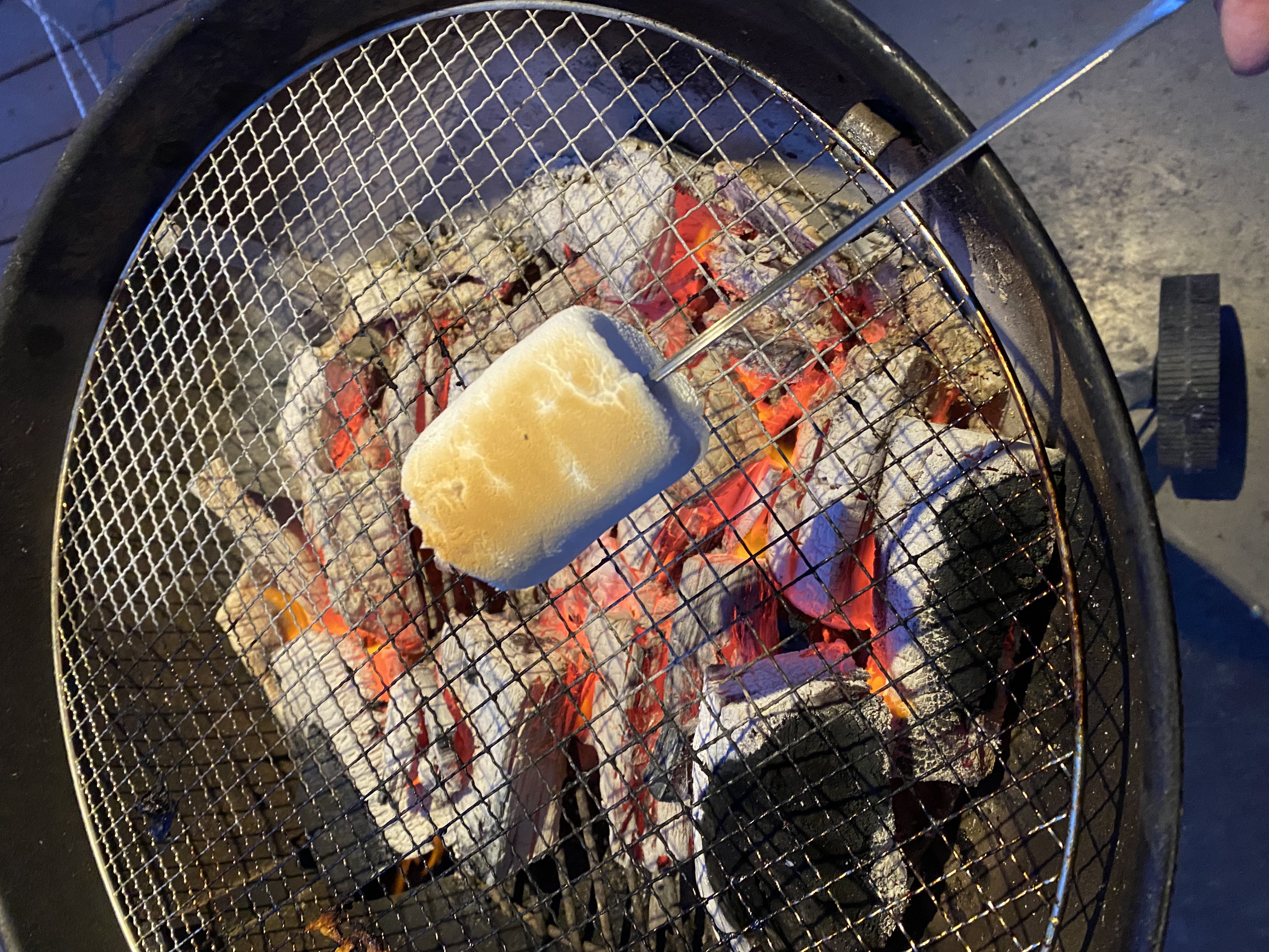 A large marshmallow being roasted on a skewer over glowing hot coals in a fire pit at night