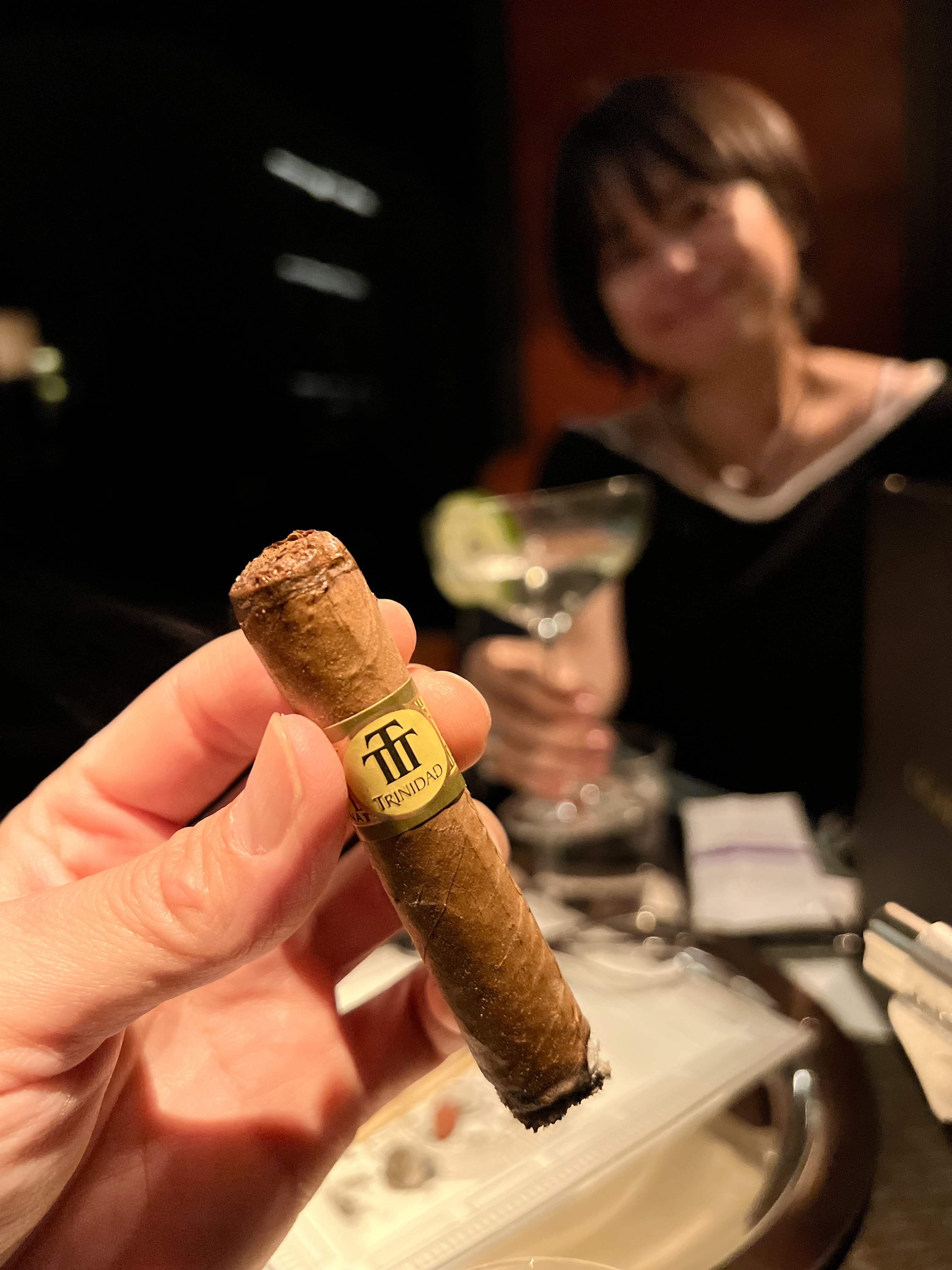 A Trinidad Reyes cigar held up mid-smoke at the Park Hyatt lounge, Ayumi visible in the background with a cocktail