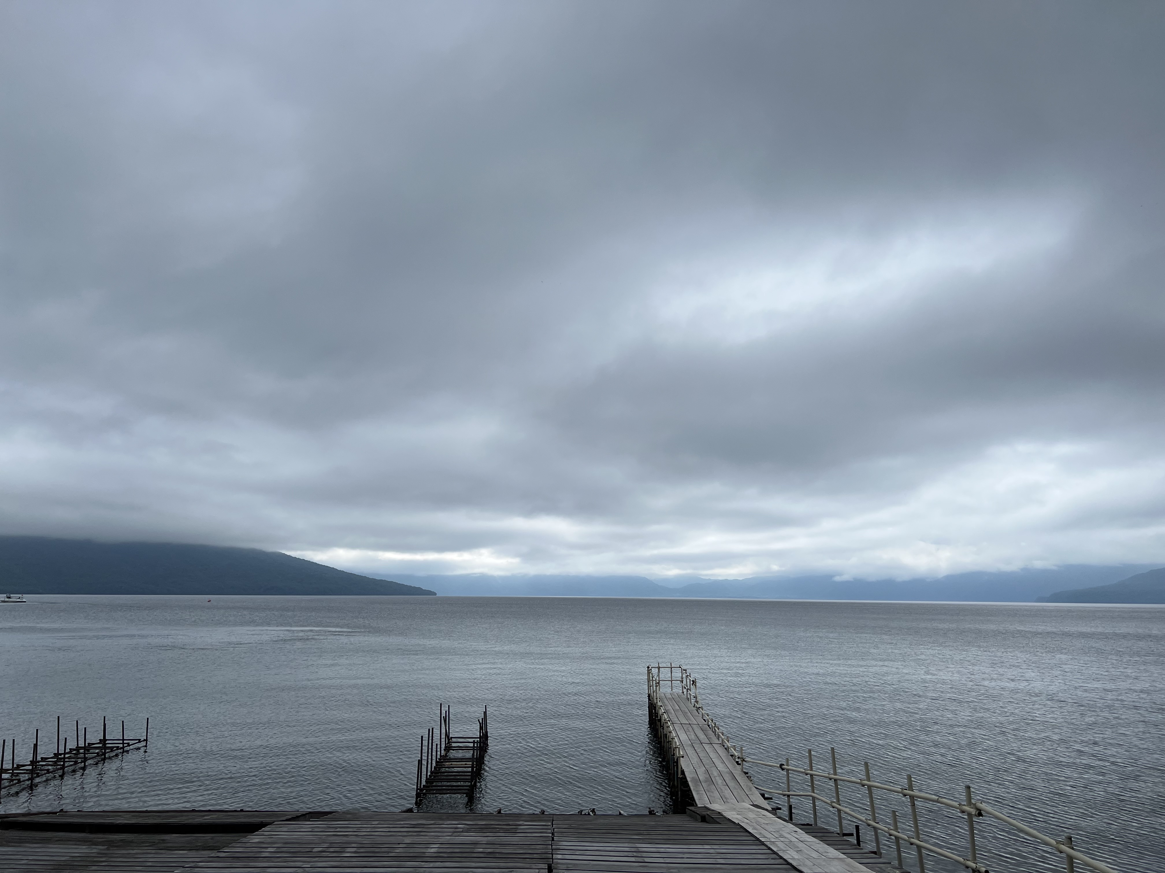 Lake Shikotsu on an overcast morning — a wooden pier extends into the still grey-blue caldera lake, mountains visible through low cloud on the far shore