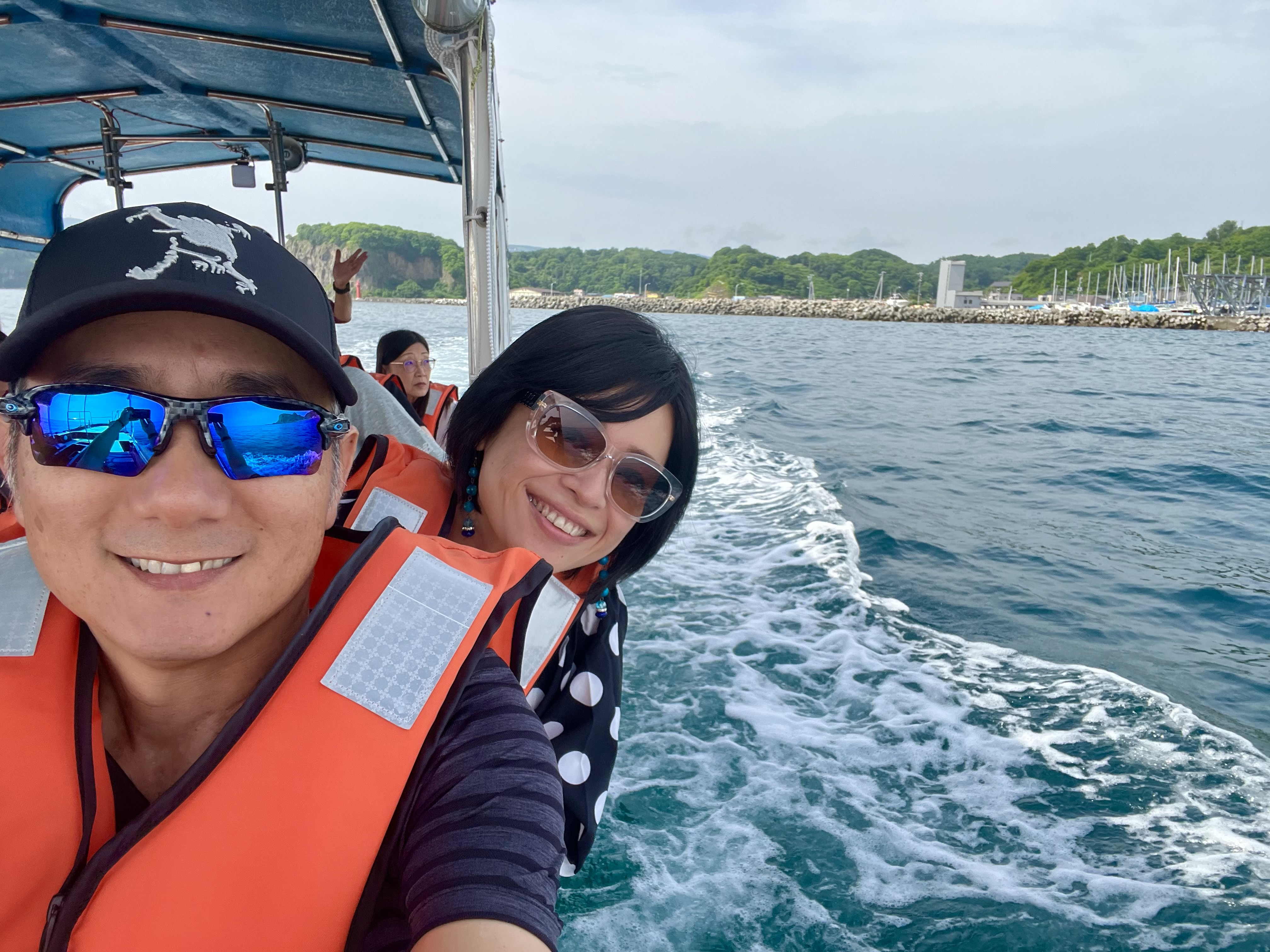 John and Ayumi on the Blue Cave cruise boat wearing orange life vests, smiling, the Otaru coast and marina visible behind them