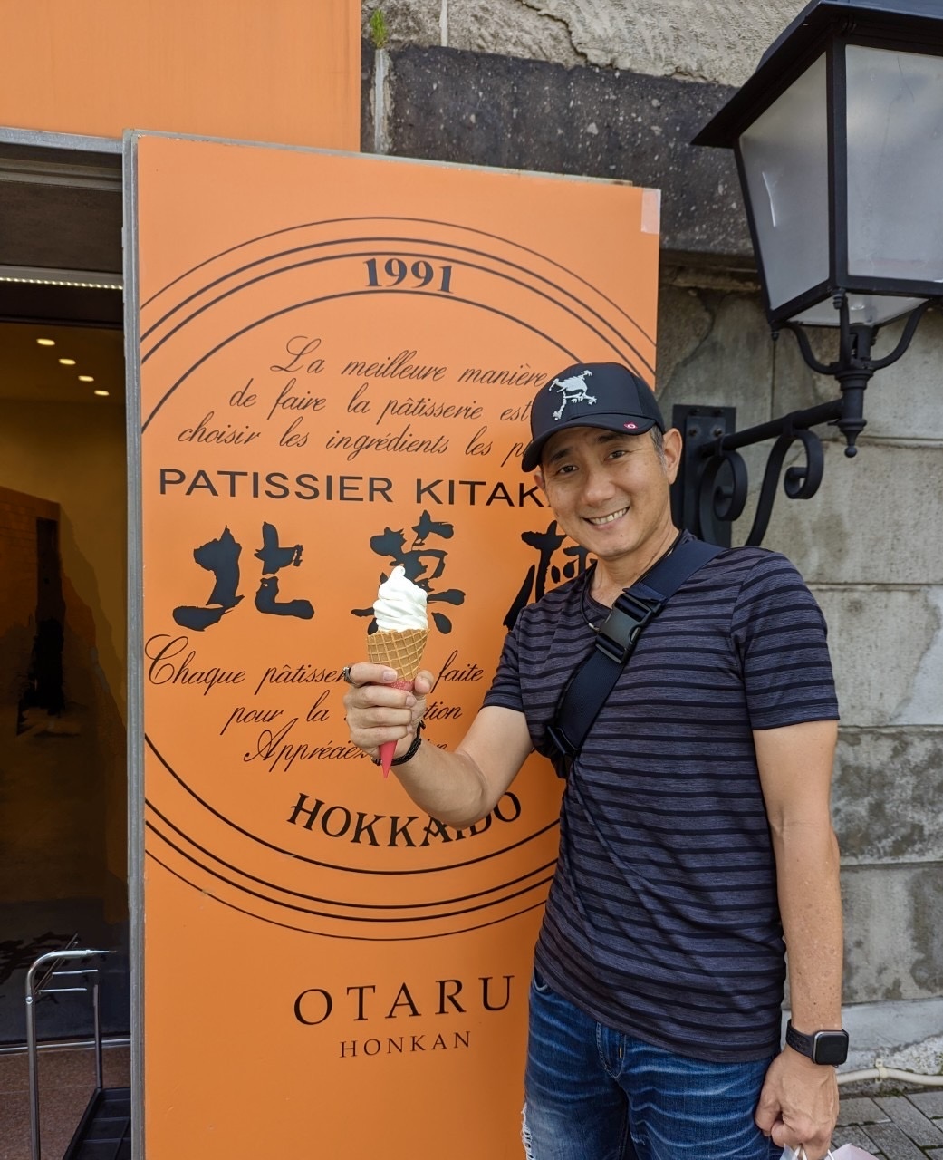 John holding a Kitakaro soft serve cone in front of the Patissier Kitakaro Otaru Honkan orange entrance sign