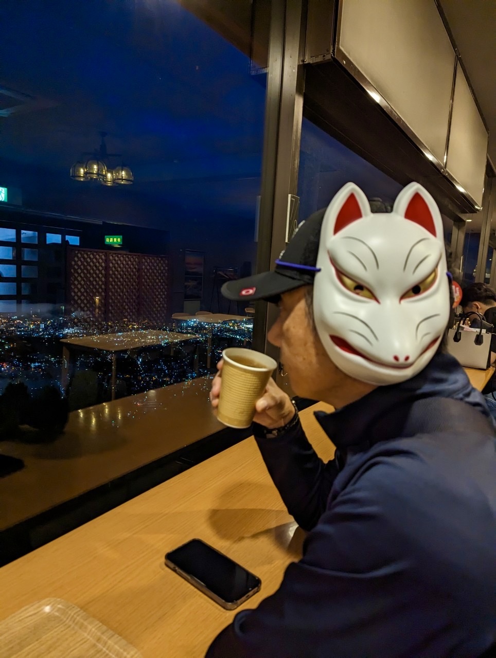 John wearing a white kitsune fox mask at Tengu Yama observation deck, Otaru city lights glowing through the window behind him